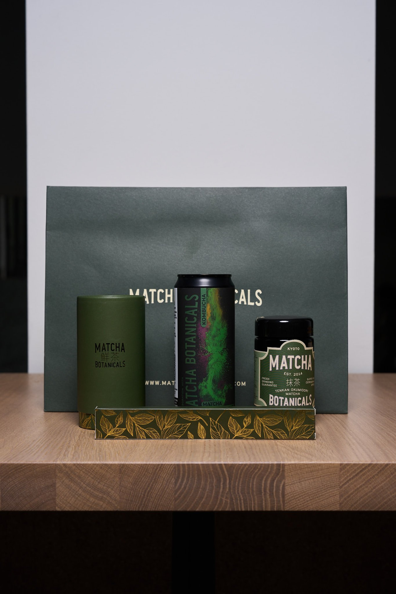 Matcha Botanicals products on a wooden table at Claw Studio: a green gift bag, a tea canister, a matcha kombucha can, a jar of matcha and a gift box